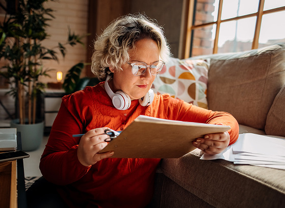 Person wearing glasses and headphones, sitting on a sofa writing on a clipboard